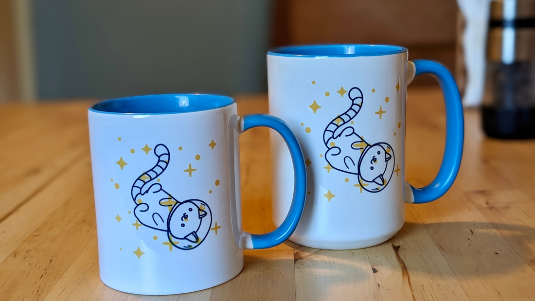 2 mugs of different sizes, both showing an upside-down cartoon kitty in a space helmet